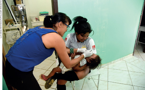 IMAGEM: duas mulheres em uma sala, uma delas está sentada segurando um bebê, enquanto a outra está aplicando uma vacina em gota na boca da criança. FIM DA IMAGEM.