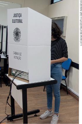 IMAGEM: mulher de pé dentro de uma cabine de papelão escrito justiça eleitoral. FIM DA IMAGEM.