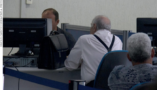 IMAGEM: um homem está ao computador prestando serviços a um idoso sentado à frente dele, há outro senhorzinho aguardando ser atendido. FIM DA IMAGEM.