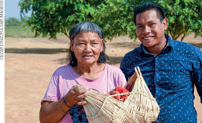 IMAGEM: homem ao lado de idosa de etnia indígena, ela segura um cesto de palha com frutas em seu interior. FIM DA IMAGEM.