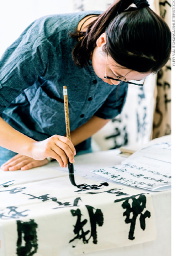 IMAGEM: garota inclinada sobre uma mesa com uma folha que cobre toda sua superfície. ela segura um pincel grande em uma das mãos e está pintando símbolos chineses com ele. FIM DA IMAGEM.