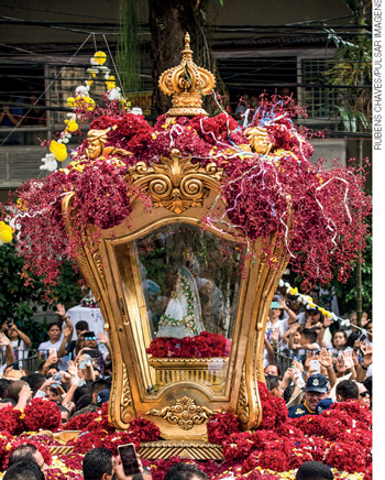 IMAGEM: a estátua de uma santa dento de uma caixa de vidro enfeitada com flores. ela é carregada entre um grande aglomerado de pessoas, que em uma festividade ao ar livre. FIM DA IMAGEM.