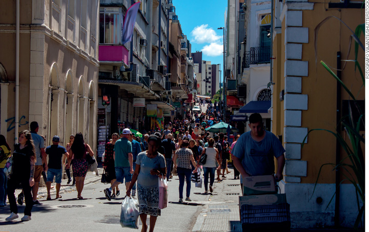 IMAGEM: rua com comércios, um grande fluxo de pessoas caminha pelo local. FIM DA IMAGEM.