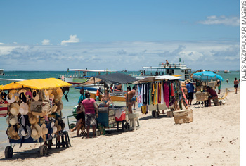 IMAGEM: barraquinhas em uma praia comercializando chapéus, alimentos, entre outros. FIM DA IMAGEM.