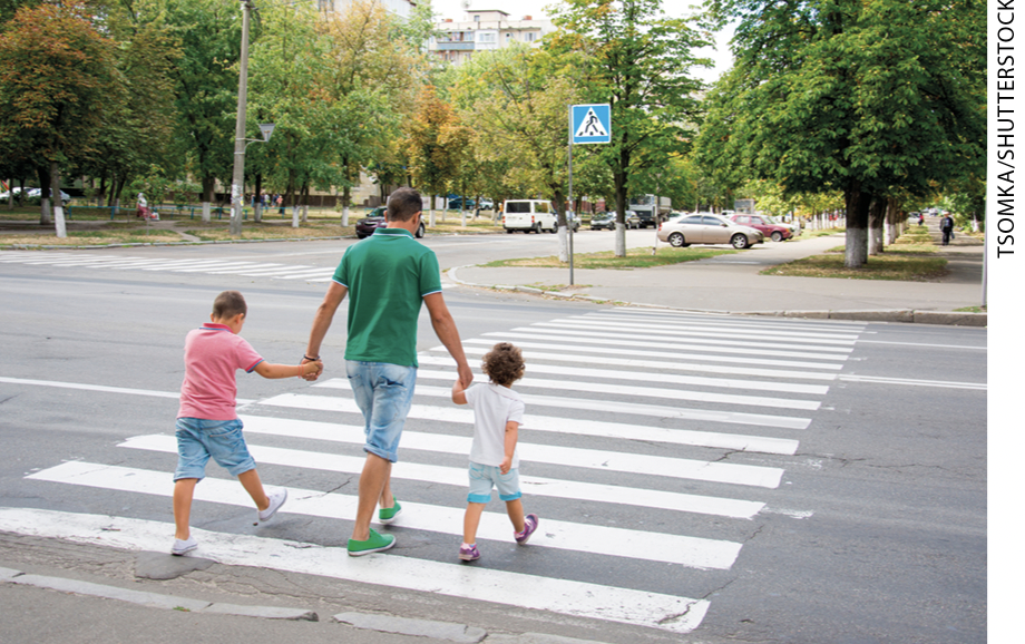 IMAGEM: homem atravessando uma faixa de pedestres segurando duas crianças pelas mãos. FIM DA IMAGEM.