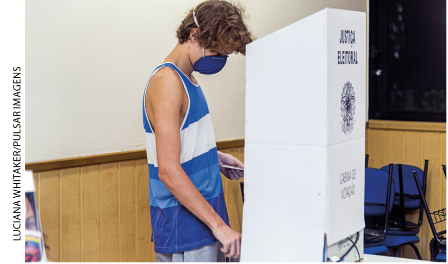 IMAGEM: jovem de pé dentro de uma cabine de papelão escrito justiça eleitoral. FIM DA IMAGEM.