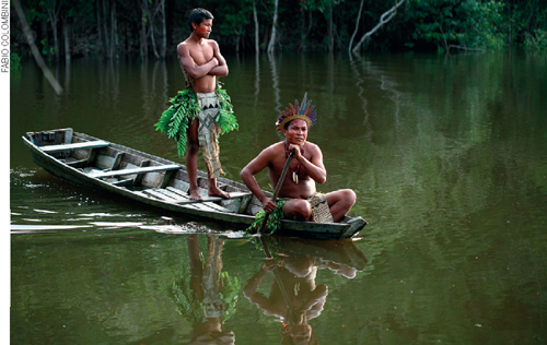 IMAGEM: dois homens indígenas em uma canoa. um está de pé logo atrás daquele que está sentado remando em um rio com vegetação densa ao fundo. FIM DA IMAGEM.