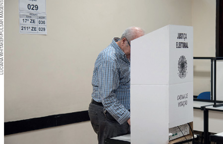 IMAGEM: homem de pé dentro de uma cabine de papelão escrito justiça eleitoral. FIM DA IMAGEM.