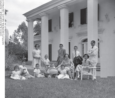IMAGEM: fotografia antiga de um grupo de pessoas reunidas em frente a frente de uma casa. FIM DA IMAGEM.