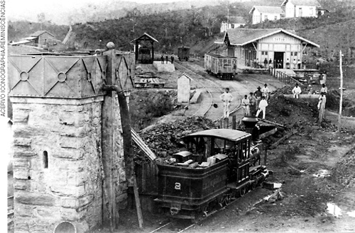 IMAGEM: fotografia antiga de uma ferrovia. há algumas construções nos arredores e um trem que passa por trilhos sobre a rua de terra batida. FIM DA IMAGEM.