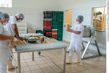 IMAGEM: três pessoas estão em um ambiente amplo, há máquinas e utensílios ao redor. os trabalhadores usam toucas e botas para manter a higiene durante o processo de fabricação de doces. FIM DA IMAGEM.