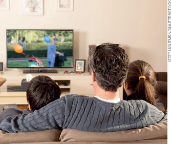 IMAGEM: família sentada em um sofá, assistindo televisão. . FIM DA IMAGEM.