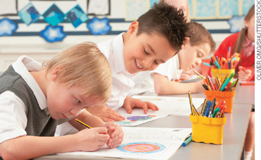 IMAGEM: crianças em uma sala de aula desenhando em folhas de papel sobre uma mesa, há alguns estojos com pincéis e lápis de cores perto delas. FIM DA IMAGEM.