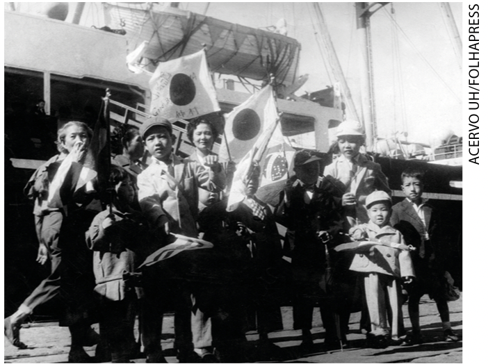 IMAGEM: fotografia antiga de pessoas comemorando à frente de um navio. FIM DA IMAGEM.