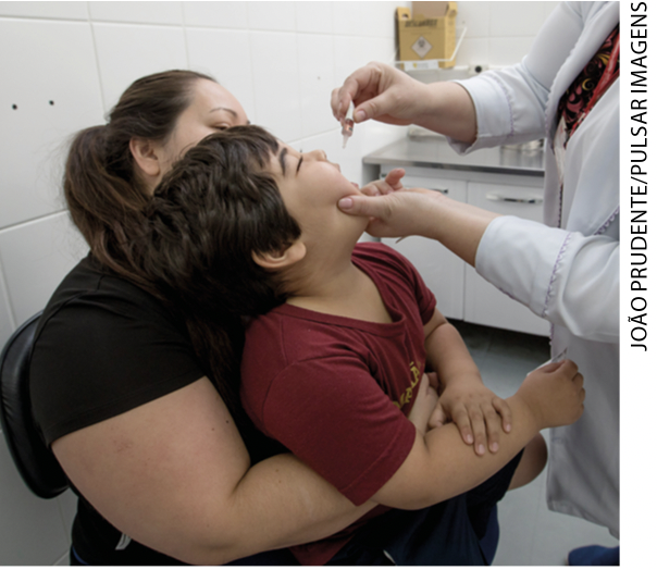 IMAGEM: criança sentada no colo de uma mulher, enquanto recebe a vacina em gotinha na boca. FIM DA IMAGEM.