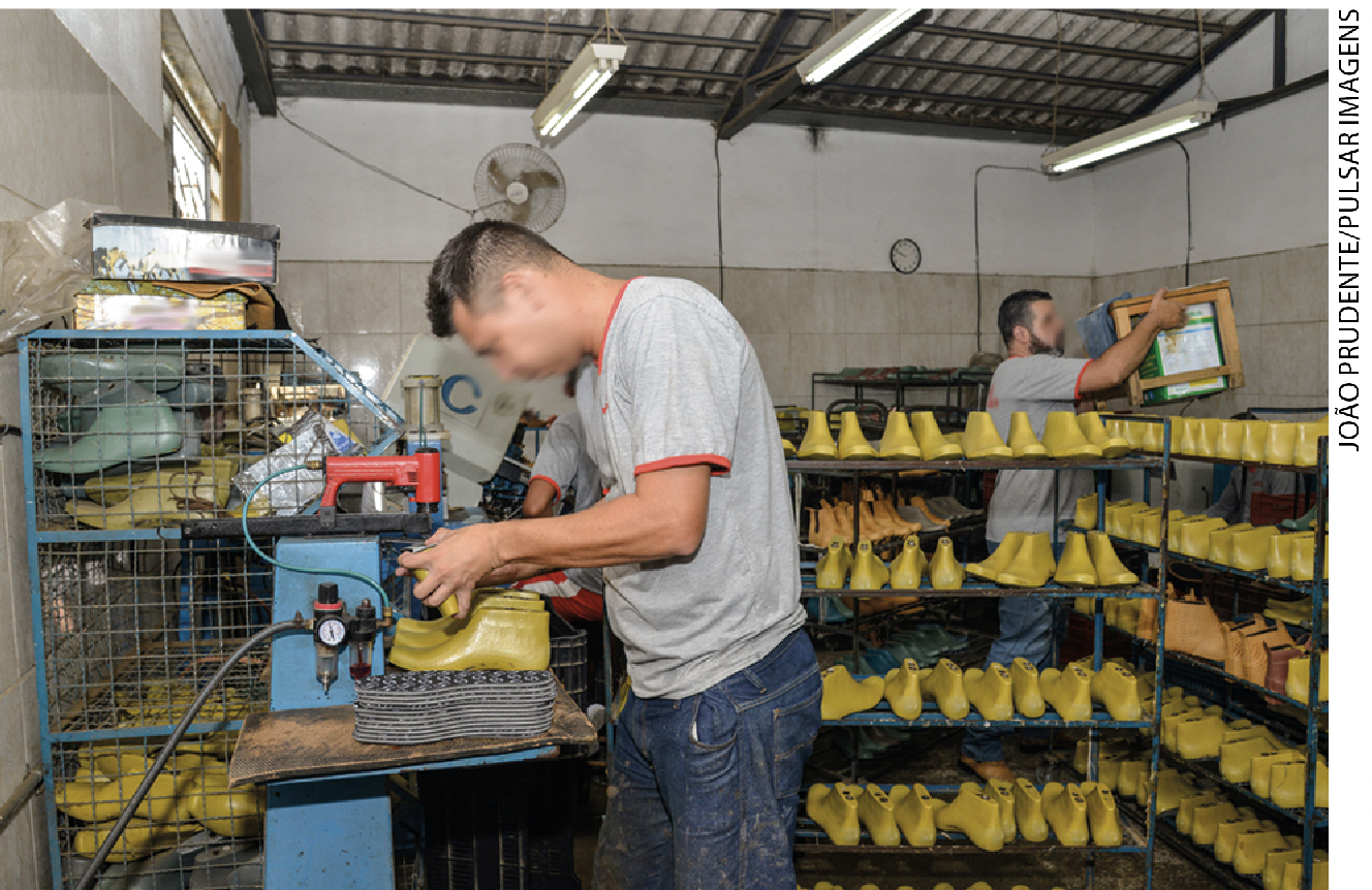 IMAGEM: trabalhadores em uma pequena fábrica de calçados, um deles manuseia um sapato em uma máquina e outro segura uma caixa nas mãos. FIM DA IMAGEM.