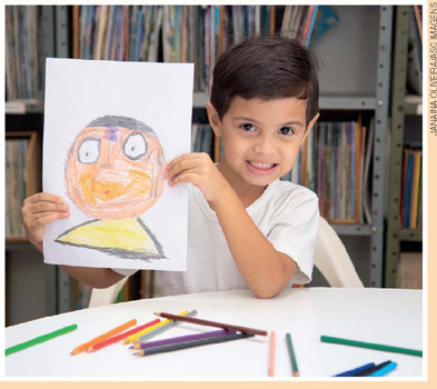 IMAGEM: um menino sorrindo, segura uma folha de sulfite com um desenho. sobre a mesa há lápis de várias cores e, ao fundo, uma estante com livros. FIM DA IMAGEM.