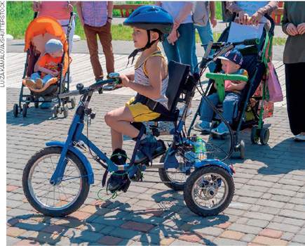 IMAGEM: fotografia de uma criança andando em um triciclo, com uma roda maior à frente e duas menores atrás. ele está sentado em uma cadeirinha e usa um capacete de proteção. ao fundo, há várias pessoas em pé e bebês em carrinhos. FIM DA IMAGEM.