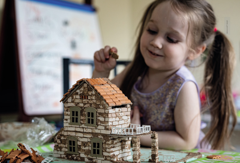 IMAGEM: uma criança utiliza peças de montar para construir uma casa com várias janelas e telhado triangular. FIM DA IMAGEM.