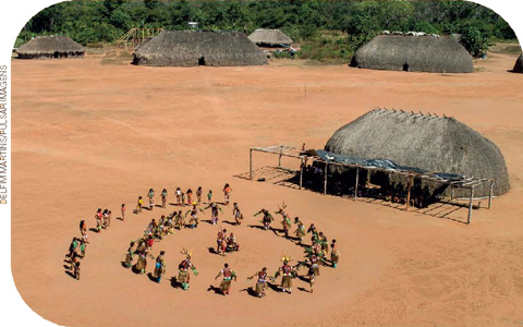 IMAGEM: vista área de uma aldeia indígena com algumas ocas espalhadas pelo terreiro. no centro do local estão várias pessoas formando uma roda. FIM DA IMAGEM.