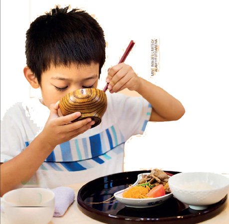 IMAGEM: um menino asiático come com dois hashis o alimento dentro de uma tigela. próximo a ele, há um prato com comidas japonesas sobre uma mesa. FIM DA IMAGEM.