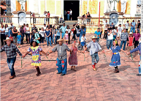 IMAGEM: adultos e idosos vestidos com roupas caipiras, dançando na praça de uma cidade em frente à igreja. ao fundo, diversas pessoas observam a apresentação. FIM DA IMAGEM.
