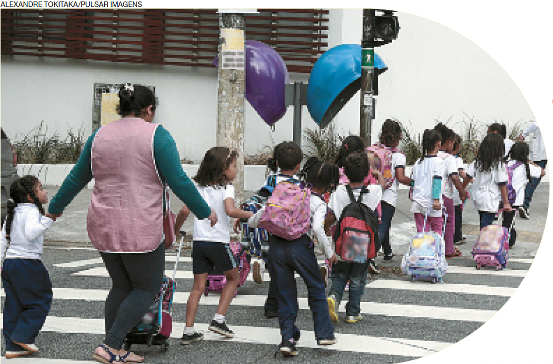 IMAGEM: várias crianças carregando mochilas nas costas e de rodinhas, atravessando a faixa de pedestres em uma rua com a ajuda de uma mulher adulta. FIM DA IMAGEM.