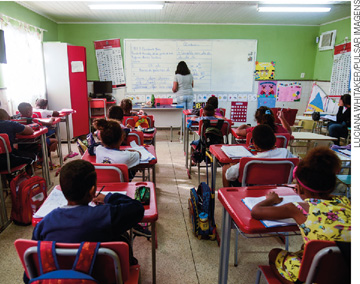 IMAGEM: crianças sentadas em carteiras escolares em uma sala de aula. todas estão de costas para o observador, assim como a professora ao fundo, que escreve na lousa. FIM DA IMAGEM.