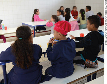 IMAGEM: crianças sentadas ao redor de grandes mesas lanchando. FIM DA IMAGEM.