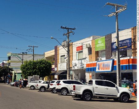 IMAGEM: rua de um bairro com carros estacionados próximos às calçadas. à direita, há uma farmácia e outros estabelecimentos comerciais. FIM DA IMAGEM.