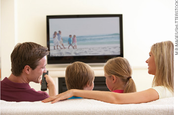 IMAGEM: no interior de uma residência, um homem, uma mulher e duas crianças assistem à televisão. FIM DA IMAGEM.