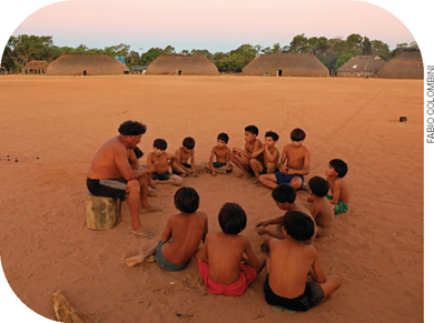 IMAGEM: várias crianças sentadas no chão de terra, em uma aldeia indígena. elas olham para um homem adulto sentado em toco de madeira. ao fundo, há várias ocas e árvores. FIM DA IMAGEM.