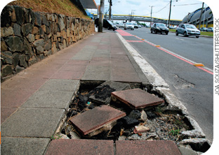 IMAGEM: uma calçada com uma parte quebrada e pedras espalhadas. ao fundo, há veículos trafegando pela rua. FIM DA IMAGEM.