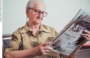IMAGEM: uma senhora de óculos lê um jornal. FIM DA IMAGEM.