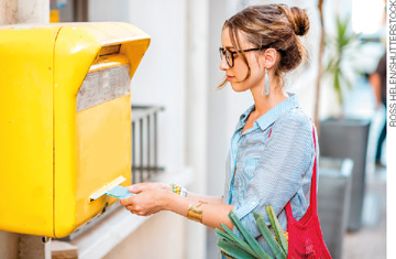 IMAGEM: uma mulher coloca uma carta em uma caixa dos correios. FIM DA IMAGEM.