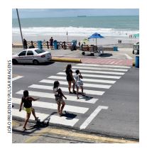 IMAGEM: pessoas atravessam a rua em uma faixa de pedestres. ao fundo, está a praia. FIM DA IMAGEM.