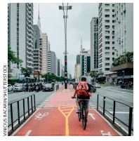 IMAGEM: uma pessoa anda de bicicleta em uma ciclovia de uma grande cidade. ao fundo, há vários prédios no entorno e carros que trafegam pela rua. FIM DA IMAGEM.