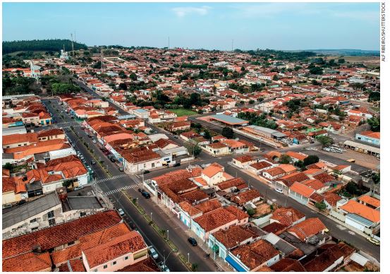 IMAGEM: vista aérea de uma cidade com vários bairros, casas, quarteirões e ruas asfaltadas. ao fundo, há montanhas e vegetação. FIM DA IMAGEM.