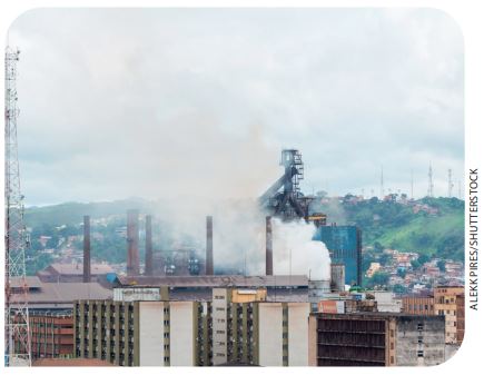 IMAGEM: uma indústria em meio a uma grande cidade, com chaminés lançando fumaça e gases poluentes na atmosfera. ao redor, há vários prédios e casas. FIM DA IMAGEM.