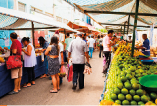 IMAGEM: 1. Pessoas compram frutas, verduras e legumes em uma feira livre de rua. À direita, há várias frutas empilhadas em uma das barracas. FIM DA IMAGEM.