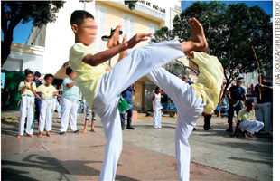 IMAGEM: dois jovens lutam capoeira no meio de uma roda formada por outros lutadores. os jovens levantam as pernas na altura da cabeça e usam calças brancas e camisetas amarelas. FIM DA IMAGEM.