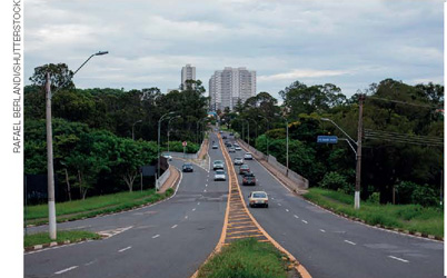 IMAGEM: duas longas avenidas asfaltadas com diversos carros e postes de iluminação cortam uma área cheia de árvores em uma cidade. FIM DA IMAGEM.