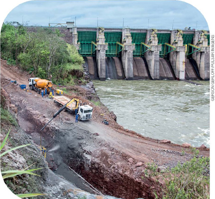 IMAGEM: caminhões e trabalhadores fazendo uma obra em uma estrada de terra na beira de um rio, onde está localizada uma usina hidrelétrica. FIM DA IMAGEM.