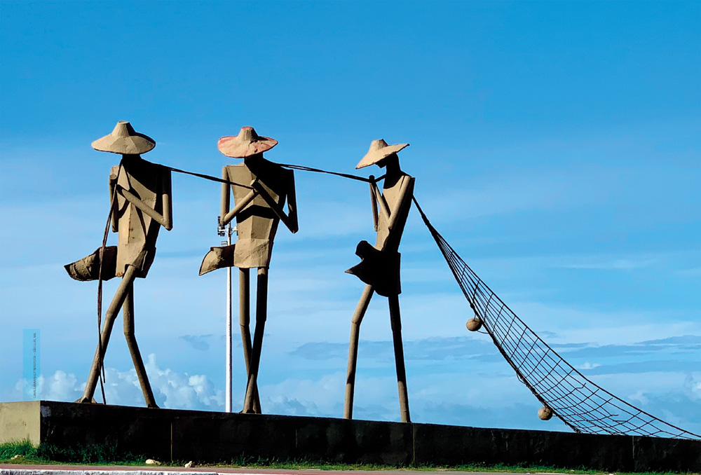 IMAGEM: a fotografia que inicia a unidade 3 mostra a escultura de três pessoas usando chapéus puxando uma rede de pesca. o monumento dos pescadores está localizado em um lugar bem alto e apenas as nuvens aparecem ao fundo. FIM DA IMAGEM.