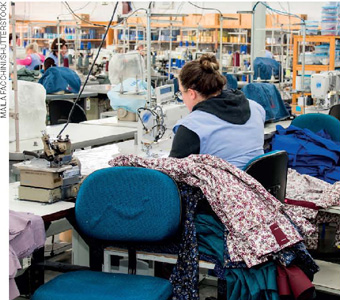 IMAGEM: uma mulher em uma indústria de vestuário está costurando com uma máquina de costura ao lado de uma pilha de roupas. FIM DA IMAGEM.