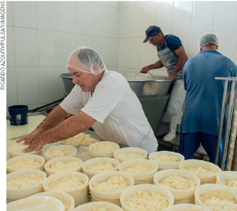 IMAGEM: um homem de avental com touca no cabelo coloca queijo em diversos recipientes. ao fundo, dois homens mexem em um grande caldeirão. FIM DA IMAGEM.