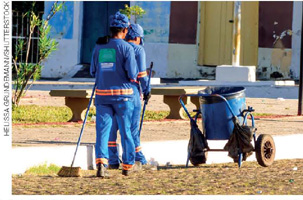 IMAGEM: garis usando uniformes e bonés fazem a limpeza de uma praça usando vassouras e uma lata de lixo. FIM DA IMAGEM.