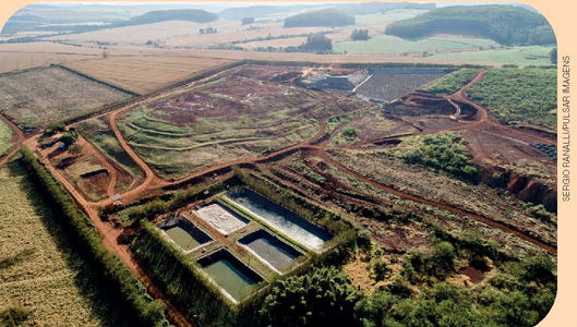 IMAGEM: vista aérea de um aterro sanitário localizado em uma planície com estradas de terra e vegetação baixa, e campos ao fundo. na parte inferior da imagem, há cinco compartimentos retangulares. FIM DA IMAGEM.