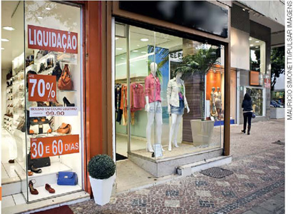 IMAGEM: vitrines de uma loja de roupa e de uma loja de sapatos com cartazes indicando liquidação dos produtos. FIM DA IMAGEM.