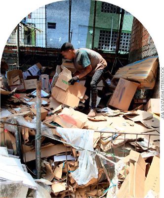 IMAGEM: pessoa separando caixas de papelão em uma pilha de lixo reciclável. FIM DA IMAGEM.